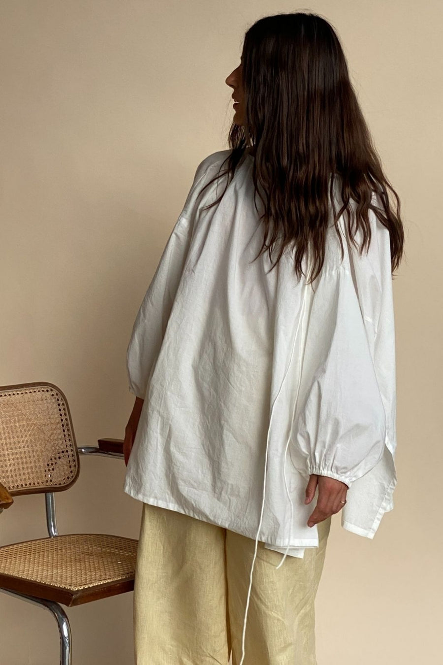 Sarah, back view, wearing the Joss blouse in White with pale yellow linen pants beside a cane chair